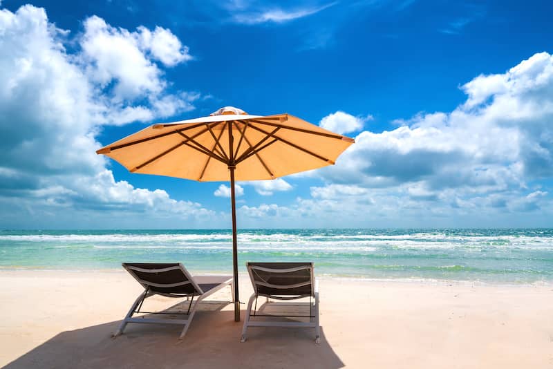 a beach sandy with blue skies and a calm sea with two deck chairs under an umbrella.