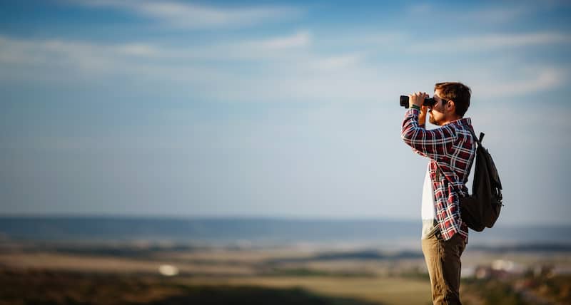 man in a checked shirt and backpack with binoculars looking out over a countryside scene.
